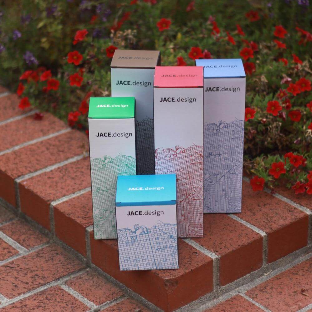 Colorful JACE.design packaging boxes arranged on outdoor bricks with vibrant flowers in the background.