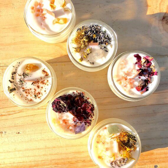 Collection of six organic intention candles with natural herbs and gemstones on wooden surface.