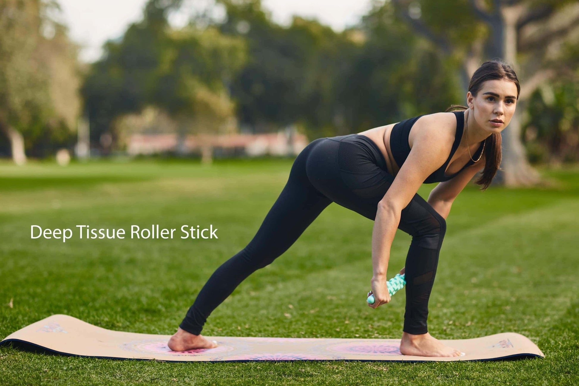 Woman using a deep tissue roller stick on a yoga mat outdoors for muscle recovery and flexibility.